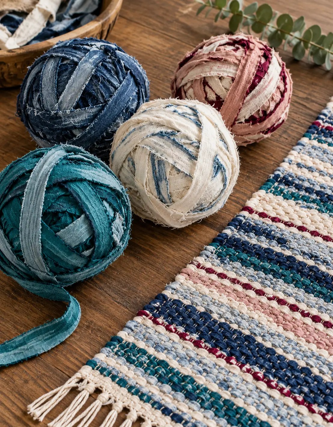 Balls of fabric-strip yarn in denim blue, teal, cream and rose beside a striped hand-woven rag rug in matching colours